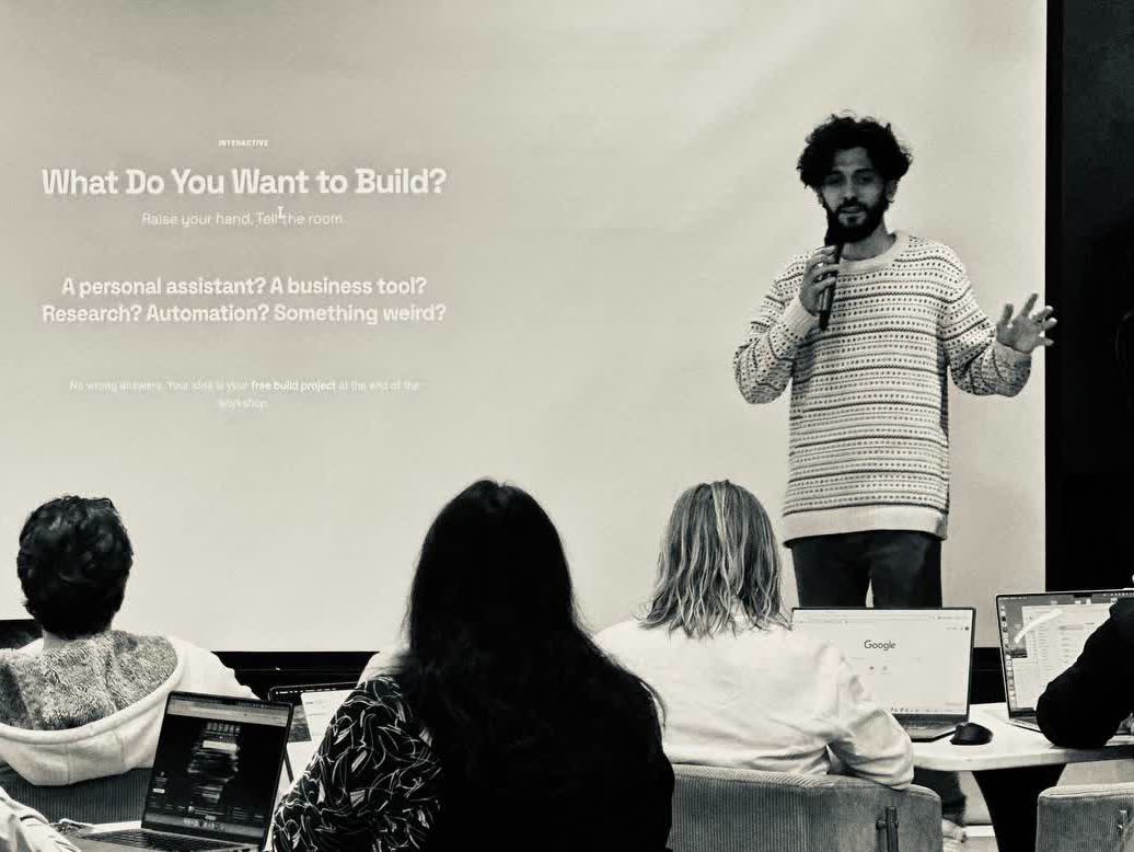 Standing beside a projected slide reading "What Do You Want to Build?" in front of a room of people working on laptops
