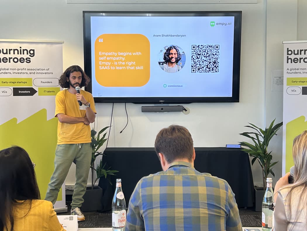 Pitching Empy to a seated audience, a slide reading "Empathy begins with self empathy" on the screen behind, Burning Heroes banners on both sides