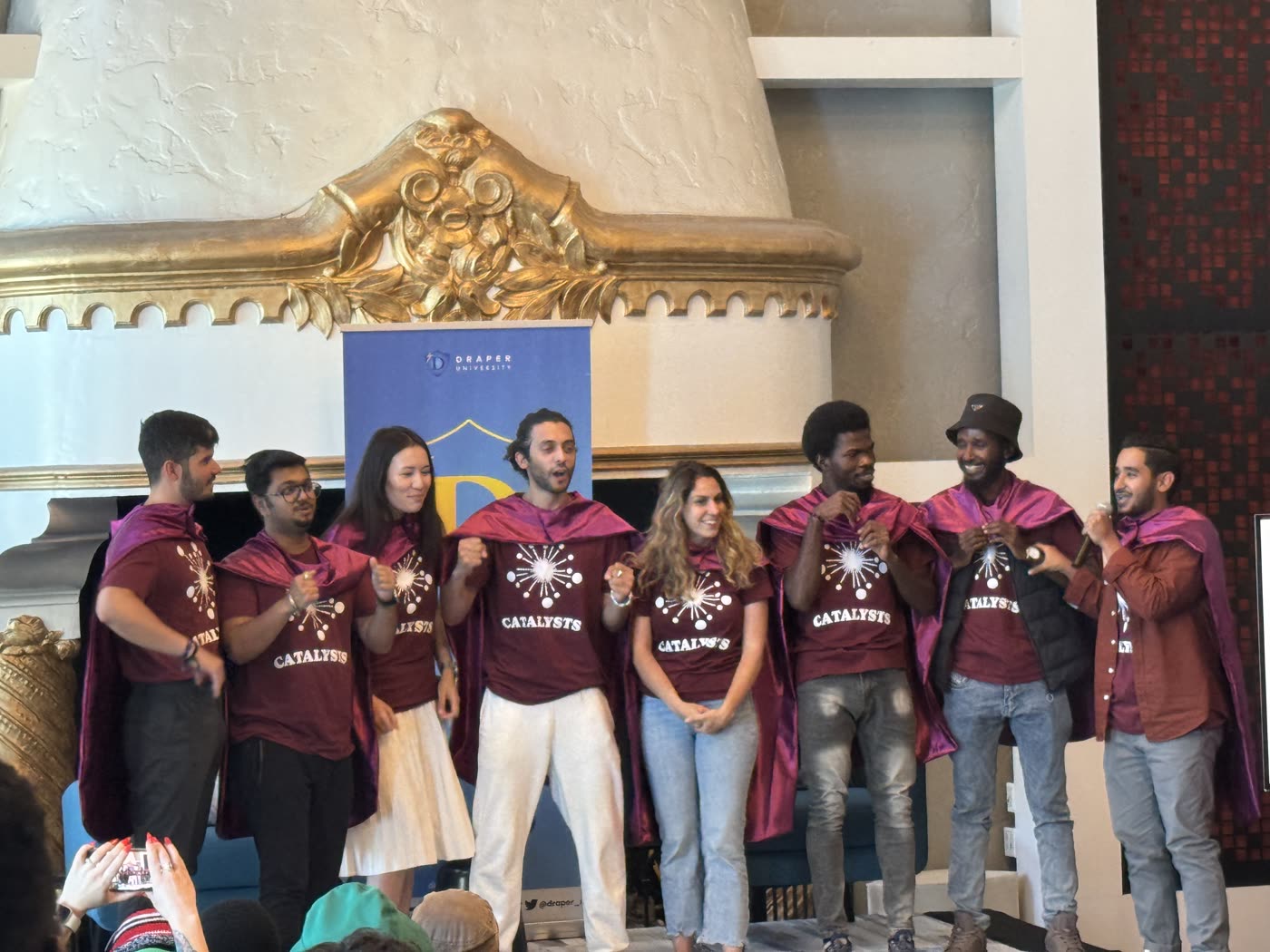 Eight people in maroon capes and Catalysts T-shirts on stage in front of a Draper University banner