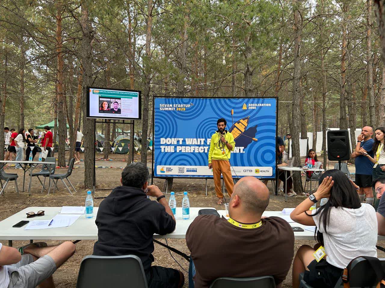 Speaking into a microphone outdoors among pine trees, in front of a Sevan Startup Summit 2025 Acceleration Zone banner, judges seated at tables in front