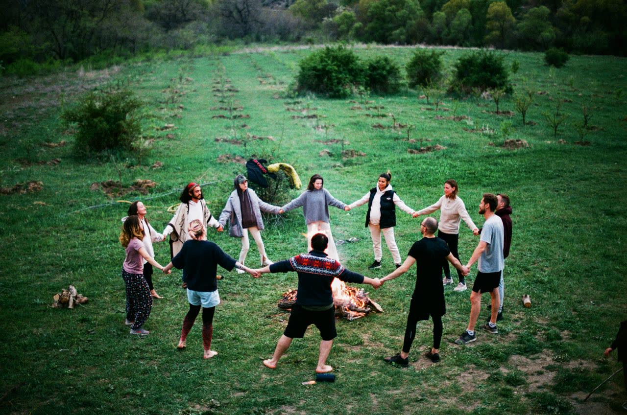 About a dozen people standing in a circle holding hands around a fire in a field at dusk