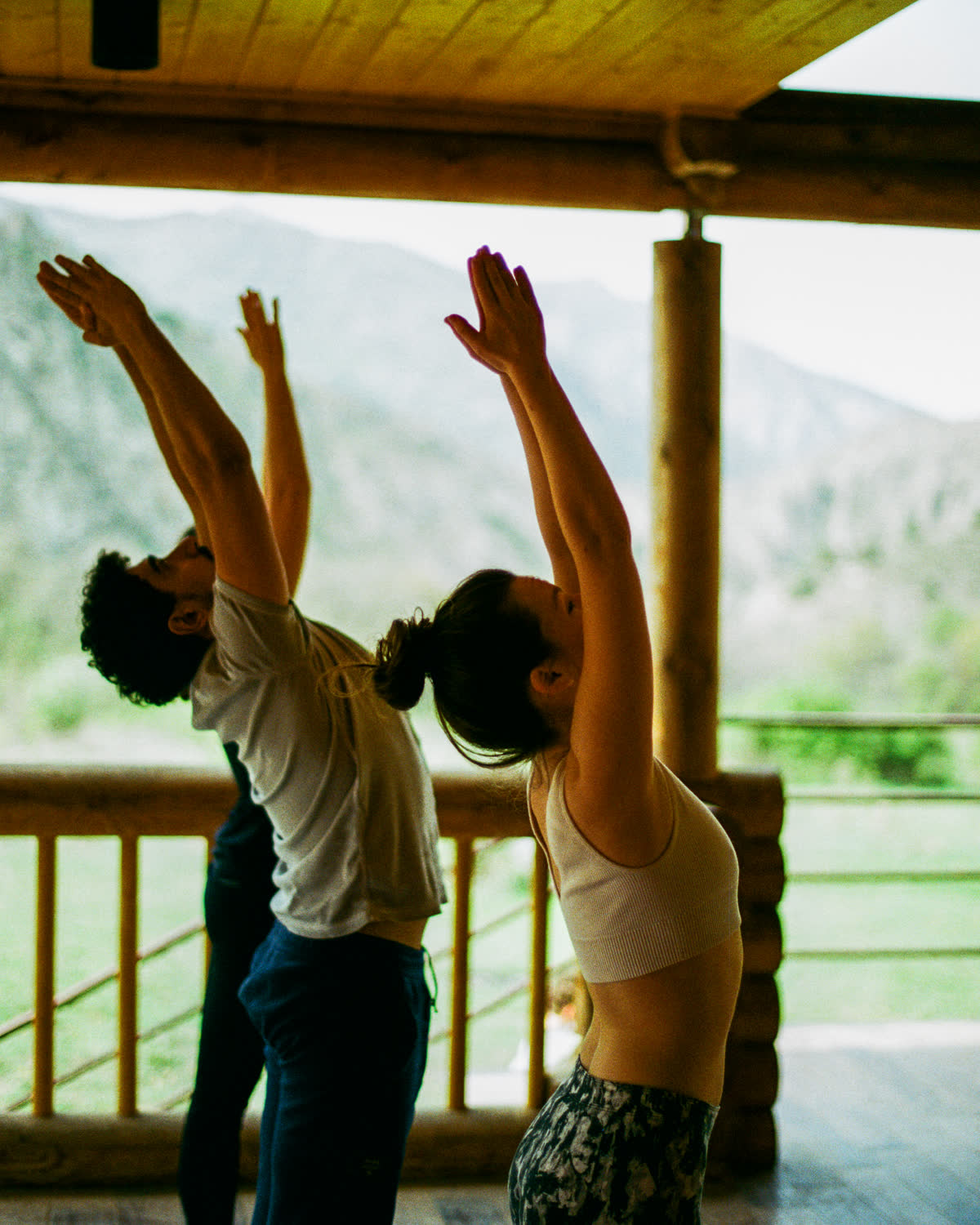 Two people on a wooden veranda in a standing backbend with arms raised, mountains behind them