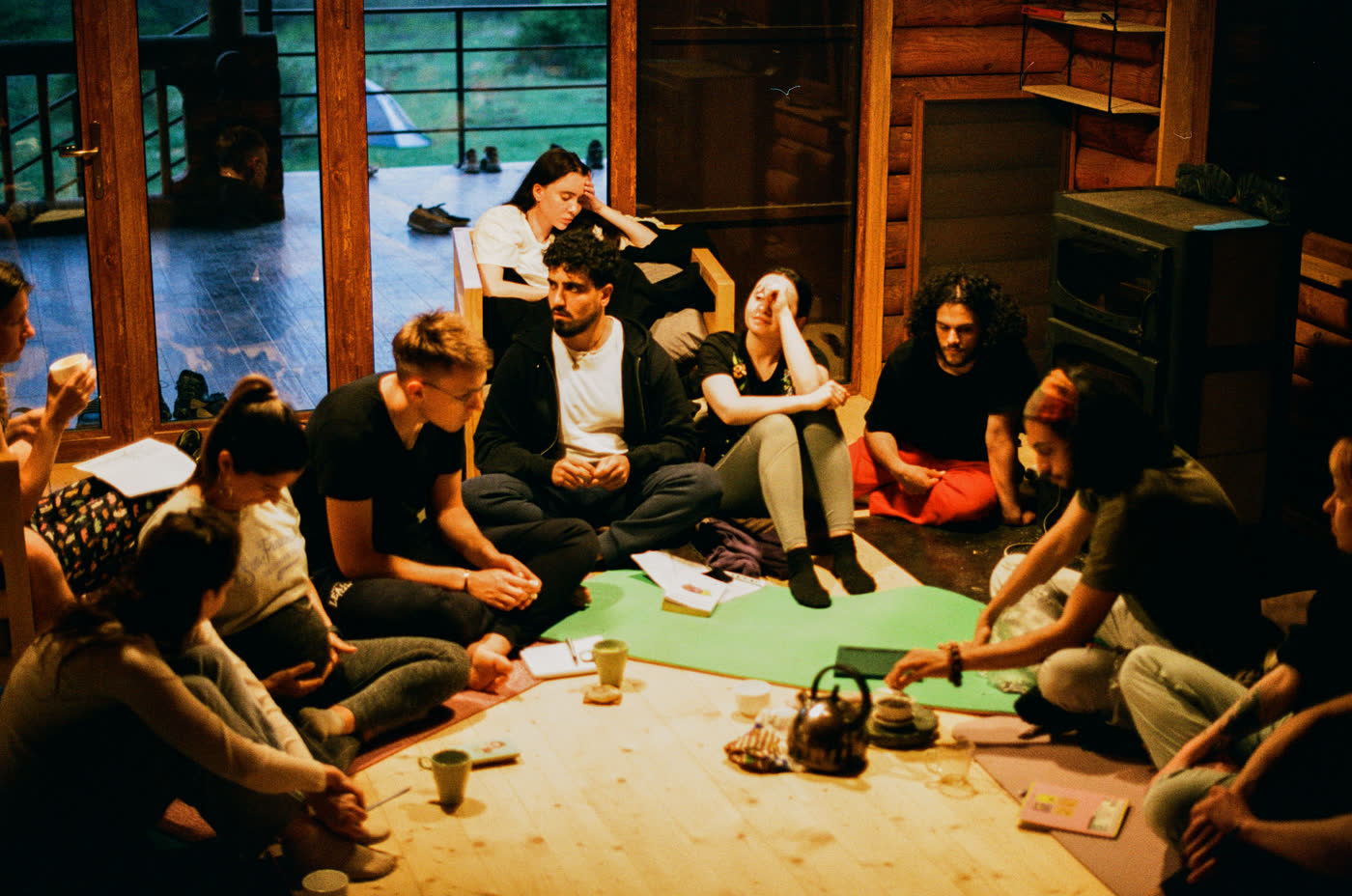 A dozen people sitting on the floor of a wooden room in a loose circle, with notebooks, mugs and a kettle between them
